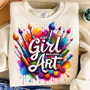 Puede incluir: Sudadera crema con el texto "Just a Girl Who Loves Art". El diseño incluye pinceles, salpicaduras de pintura y una paleta de colores vibrantes, ideal para los amantes del arte.