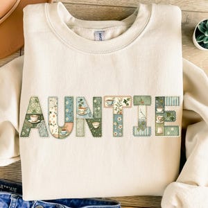 Puede incluir: Sudadera color crema con la palabra "AUNTIE" en letras de patchwork. Cada letra presenta un diseño floral o estampado diferente, con tazas de té incorporadas. La sudadera tiene cuello redondo y mangas largas.