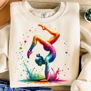 Può includere: Una felpa bianca con una grafica colorata di una ginnasta che esegue un handstand. La ginnasta indossa un body rosa ed è circondata da schizzi di vernice colorati.