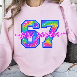 K&ouml;nnte beinhalten: Ein hellrosa Sweatshirt mit der Zahl "67" in einem lebendigen, regenbogenfarbenen Design mit blauem Umriss. Die Worte "six seven" sind in einer kursiven Schrift &uuml;ber den Zahlen geschrieben. Das Sweatshirt ist ein l&auml;ssiger Stil.