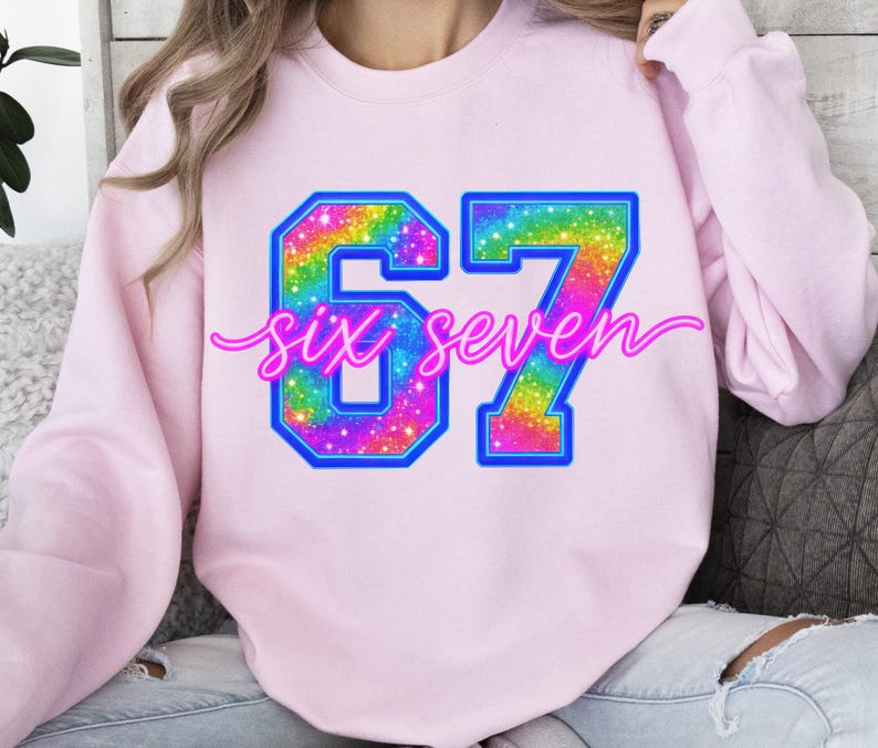 K&ouml;nnte beinhalten: Ein hellrosa Sweatshirt mit der Zahl "67" in einem lebendigen, regenbogenfarbenen Design mit blauem Umriss. Die Worte "six seven" sind in einer kursiven Schrift &uuml;ber den Zahlen geschrieben. Das Sweatshirt ist ein l&auml;ssiger Stil.