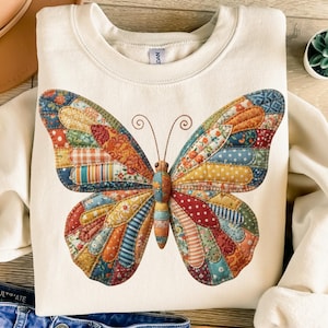 May include: Cream-colored sweatshirt featuring a large, colorful butterfly design. The butterfly wings are composed of various fabric patches in shades of red, orange, blue, and yellow. The butterfly has brown antennae and a detailed body. The sweatshirt is made of a soft material.