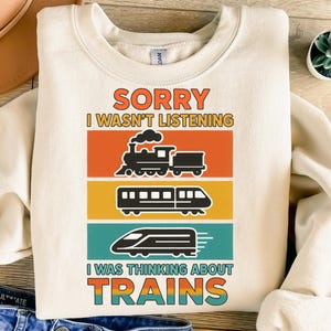 Peut inclure: Sweat-shirt crème avec le texte "SORRY I WASN'T LISTENING I WAS THINKING ABOUT TRAINS." Le design présente trois illustrations de trains dans des rectangles orange, jaune et turquoise.