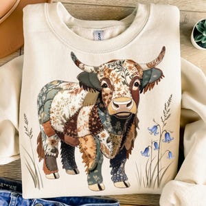 May include: Cream-colored sweatshirt featuring a patchwork-style Highland cow design. The cow has a detailed pattern of floral and geometric shapes in various colors. Bluebell flowers and tall grass are also depicted.