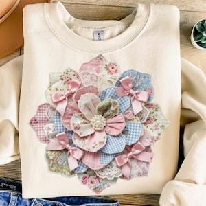 Può includere: Felpa color crema con un'ampia applicazione floreale. L'applicazione è realizzata con vari tessuti fantasia in rosa, blu e verde, con fiocchi di raso rosa e un fiore con perle al centro. La felpa ha uno scollo tondo.
