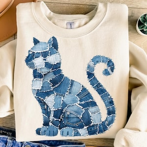 Könnte beinhalten: Cremefarbenes Sweatshirt mit einem Katzenmotiv aus Jeansstoff. Die Katze besteht aus verschiedenen Jeansstoff-Flicken in Blautönen. Die Katze sitzt mit eingerolltem Schwanz. Das Sweatshirt hat einen Rundhalsausschnitt und lange Ärmel.