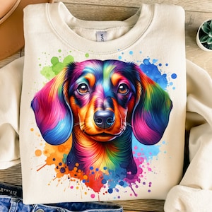 Könnte beinhalten: Ein weißer Pullover mit einer bunten Illustration eines Dackels. Der Hund ist in einem Regenbogen aus Farben mit Aquarellspritzer im Hintergrund gemalt.