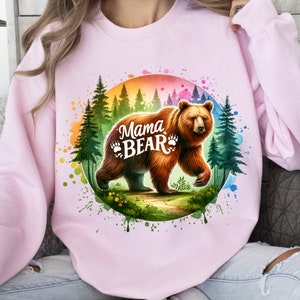 May include: Light pink sweatshirt featuring a watercolor illustration of a brown bear with the text "Mama Bear" and paw prints. The bear is set against a backdrop of a forest and a colorful rainbow circle.