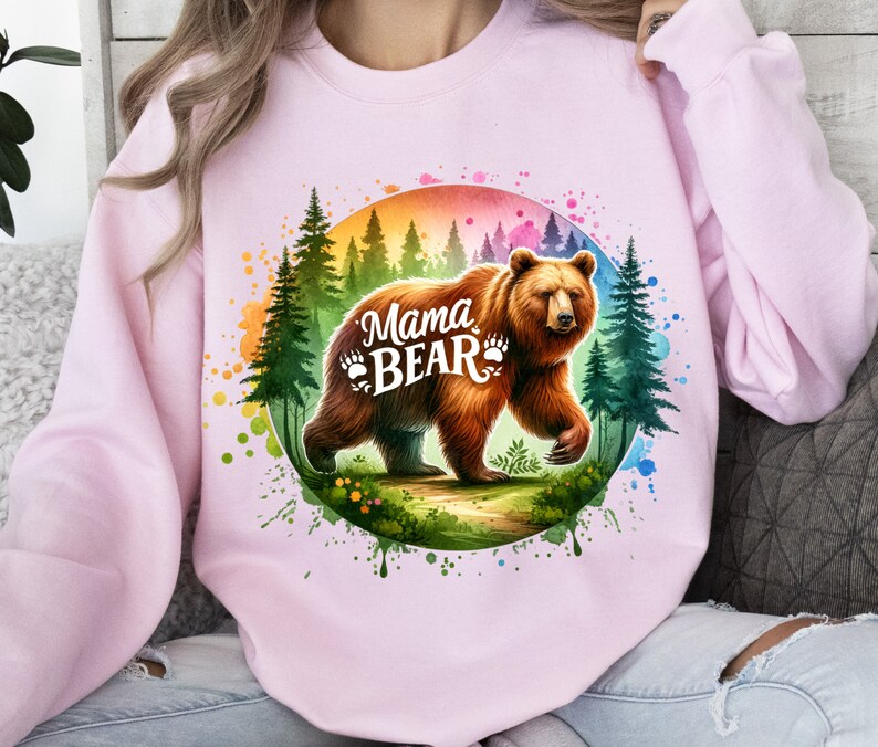 May include: Light pink sweatshirt featuring a watercolor illustration of a brown bear with the text "Mama Bear" and paw prints. The bear is set against a backdrop of a forest and a colorful rainbow circle.