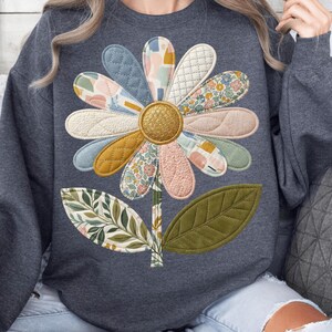 Puede incluir: Una sudadera gris oscuro con un gran aplique floral colorido. La flor tiene pétalos estampados en varios tonos de azul, rosa, amarillo y verde, con un centro dorado. El tallo y las hojas también están aplicados.