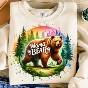 May include: Cream-colored sweatshirt featuring a watercolor illustration of a brown bear with the text "Mama Bear." The bear is set against a backdrop of a forest and a colorful rainbow-like circle. The design includes bear paw prints.