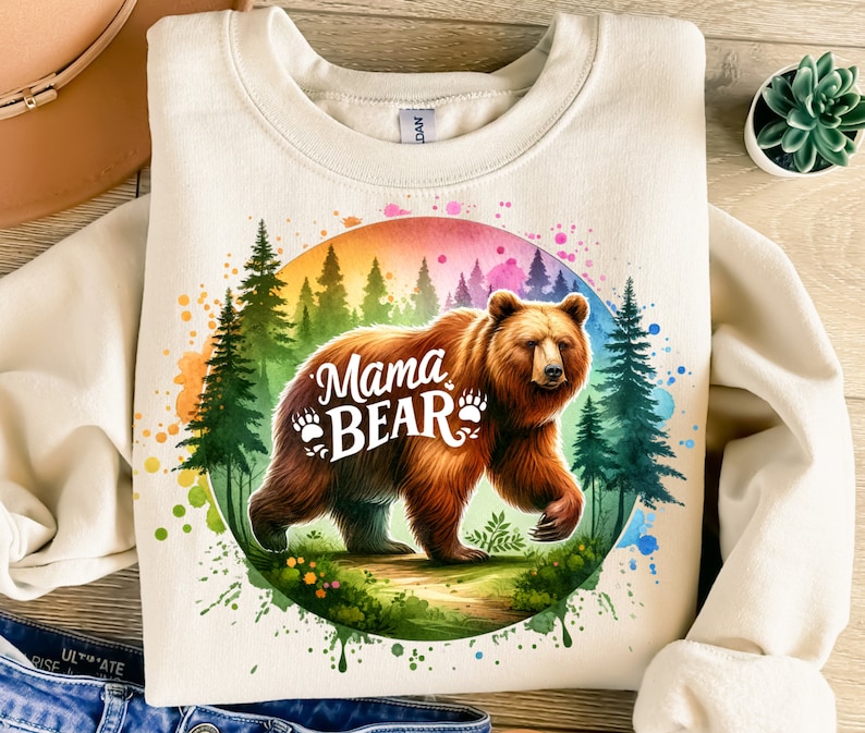 May include: Cream-colored sweatshirt featuring a watercolor illustration of a brown bear with the text "Mama Bear." The bear is set against a backdrop of a forest and a colorful rainbow-like circle. The design includes bear paw prints.