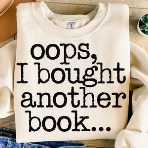 Könnte beinhalten: Ein cremefarbener Pullover mit dem Aufdruck "oops, I bought another book..." in Schwarz.