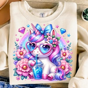 May include: Cream-coloured sweatshirt with a vibrant unicorn design. The unicorn has pink and purple hair, heart-shaped glasses, and is surrounded by flowers, gems, and a rainbow. It is holding a drink with ice cubes.