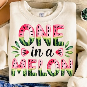 Pode incluir: Uma camisa de moletom branca com o texto "One in a Melon" em letras pretas. As letras são decoradas com fatias de melancia.