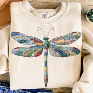 May include: Cream-colored sweatshirt featuring a large, colorful dragonfly design. The dragonfly's wings are patchwork with floral and patterned fabrics in shades of blue, red, and green. The body is striped and segmented. A casual, nature-themed top.