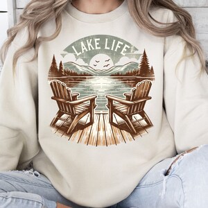May include: A beige sweatshirt featuring a lakeside scene with two wooden Adirondack chairs on a dock. The graphic includes a lake, mountains, trees, and the text "LAKE LIFE" in a circular design. The design is in shades of brown, green, and blue.