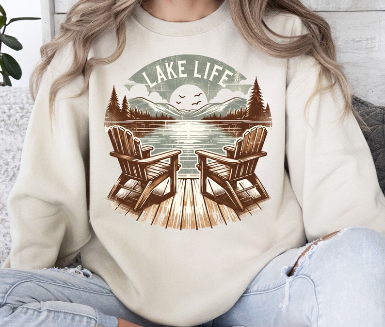 May include: A beige sweatshirt featuring a lakeside scene with two wooden Adirondack chairs on a dock. The graphic includes a lake, mountains, trees, and the text "LAKE LIFE" in a circular design. The design is in shades of brown, green, and blue.