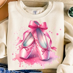 May include: A light beige sweatshirt featuring a pink watercolour design of ballet shoes with a pink bow. The shoes are on a white background with pink splatters.