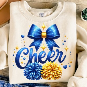 Peut inclure: Un sweat-shirt blanc avec un nœud de pom-pom de cheerleader bleu et or pailleté, des pompons et le mot "Cheer" en lettres pailletées.