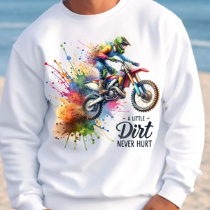 May include: White sweatshirt featuring a colorful watercolor design of a motocross rider on a red and blue dirt bike. The text "A Little Dirt Never Hurt" is below the image. The design includes splashes of color.