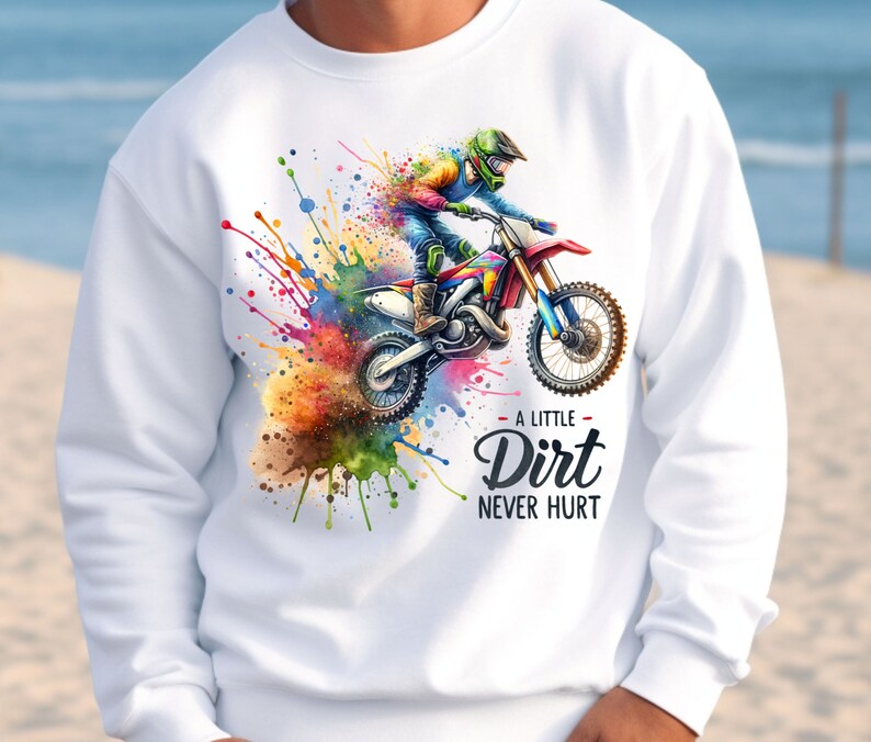 May include: White sweatshirt featuring a colorful watercolor design of a motocross rider on a red and blue dirt bike. The text "A Little Dirt Never Hurt" is below the image. The design includes splashes of color.