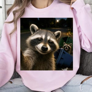 May include: A light pink sweatshirt featuring a square image of two raccoons. One raccoon is taking a selfie, while the other is in the background near a green and blue trash bin. The background shows a street at night.