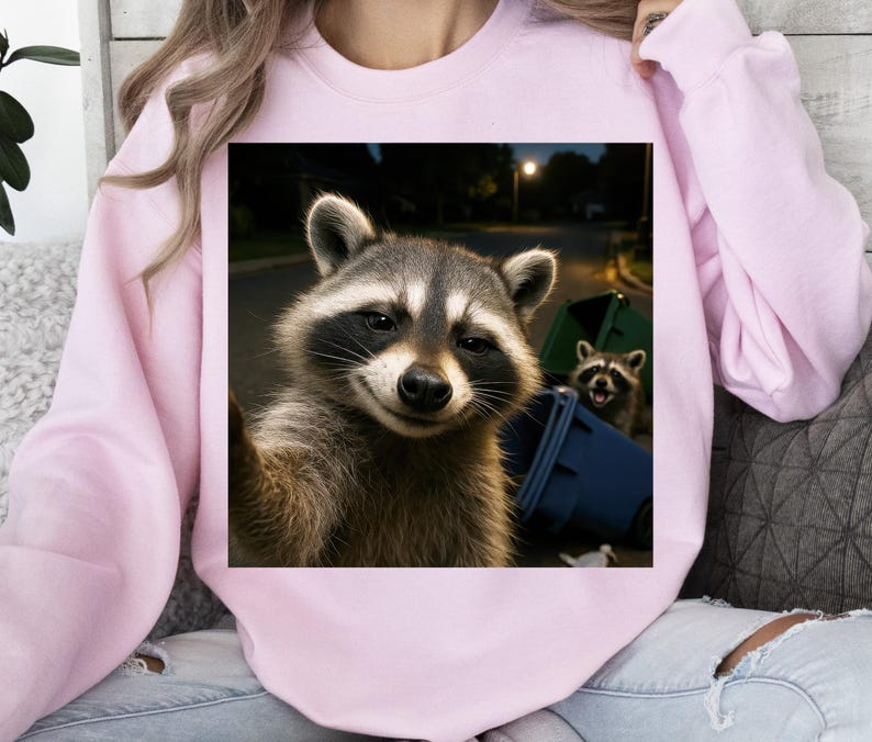 May include: A light pink sweatshirt featuring a square image of two raccoons. One raccoon is taking a selfie, while the other is in the background near a green and blue trash bin. The background shows a street at night.