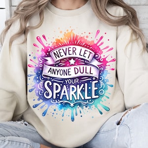May include: A cream coloured sweatshirt with a watercolour design of colourful paint splatters. The design features the text "Never let anyone dull your sparkle".