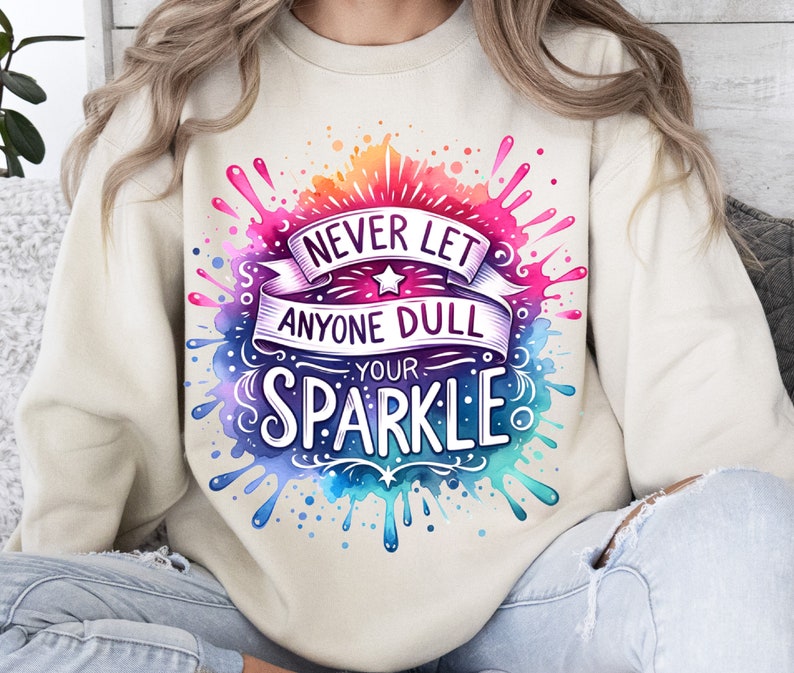 May include: A cream coloured sweatshirt with a watercolour design of colourful paint splatters. The design features the text "Never let anyone dull your sparkle".