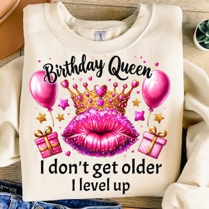 May include: A cream-colored sweatshirt with the text "Birthday Queen I don't get older I level up" in black. The design features a pink crown with gold accents, pink lips, and pink balloons.