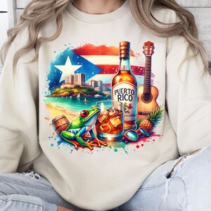 May include: A white sweatshirt with a colourful design featuring a bottle of Puerto Rican rum, a guitar, a frog, and a glass of rum. The design also includes a pineapple, a barrel, and a watercolour-style background.