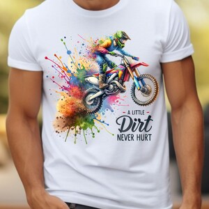 May include: White t-shirt featuring a colorful watercolor illustration of a motocross rider on a dirt bike. The design includes vibrant paint splatters and the text "A Little Dirt Never Hurt."