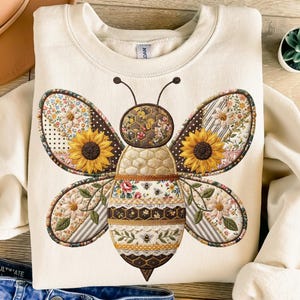 May include: Cream-colored sweatshirt featuring a large, detailed bee design. The bee has patchwork wings with sunflowers and floral patterns. The body of the bee is a mix of textures and patterns, including honeycombs and lace. A unique and stylish design.