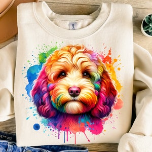 Puede incluir: Una sudadera blanca con una ilustración acuarela colorida de la cara de un perro. El perro tiene el pelo marrón y está rodeado de salpicaduras de rosa, azul, amarillo, verde y naranja.