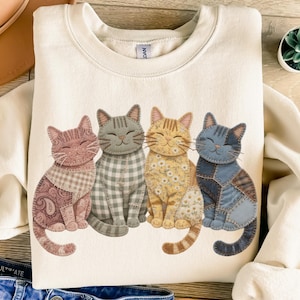 May include: Cream-coloured sweatshirt with four smiling cat illustrations. Each cat has a unique patchwork design in shades of pink, grey, yellow, and blue. The cats are arranged side by side, creating a whimsical and cosy design.