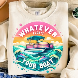 May include: A beige sweatshirt with a colorful graphic design featuring a boat, dolphins, and the text "Whatever Floats Your Boat".