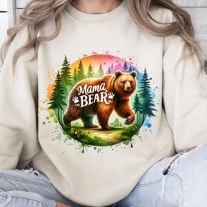 May include: Beige sweatshirt featuring a watercolor graphic of a brown bear walking through a forest. The text "Mama Bear" is displayed in white, with paw prints. The design includes a rainbow-colored circle and green trees.