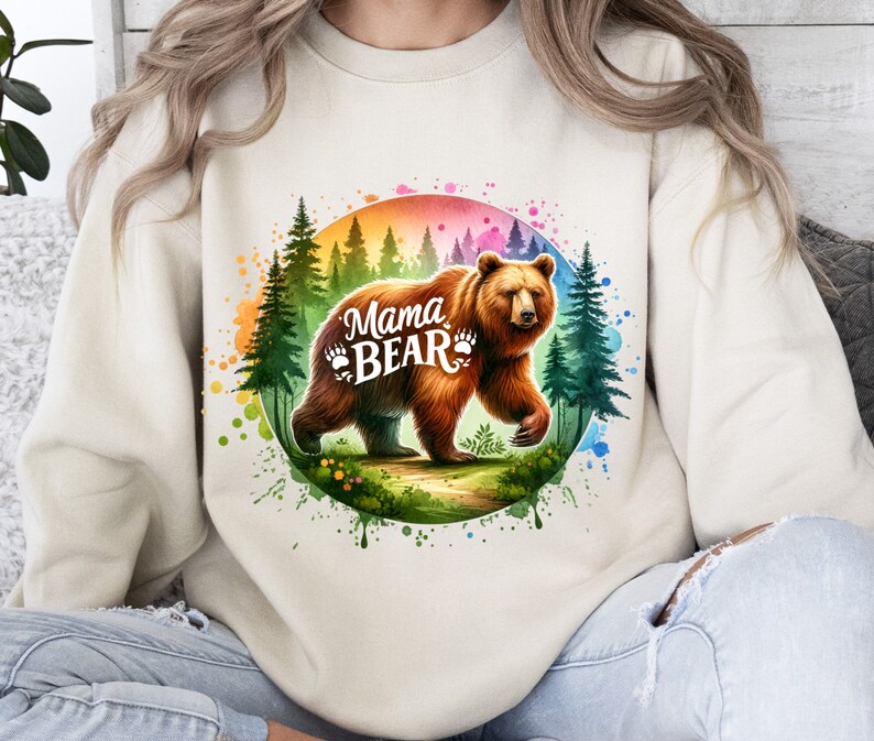 May include: Beige sweatshirt featuring a watercolor graphic of a brown bear walking through a forest. The text "Mama Bear" is displayed in white, with paw prints. The design includes a rainbow-colored circle and green trees.