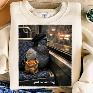 May include: Cream-colored sweatshirt featuring a framed image of a pigeon wearing a beret, looking out a window. The image includes a paper bag with a croissant and the text "just commuting".