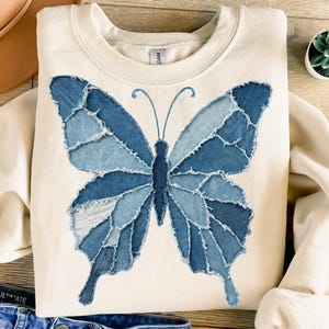 May include: A cream-coloured sweatshirt with a large butterfly design made from various shades of blue denim. The butterfly has detailed wings and a central body, creating a unique, textured look. The sweatshirt is laid flat on a wooden surface.
