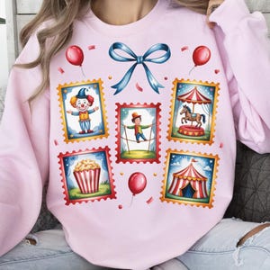 May include: A light pink sweatshirt featuring a circus-themed design. The design includes postage stamp-style illustrations of a clown, a tightrope walker, a carousel, popcorn, and a circus tent. Red balloons and a blue bow complete the design.