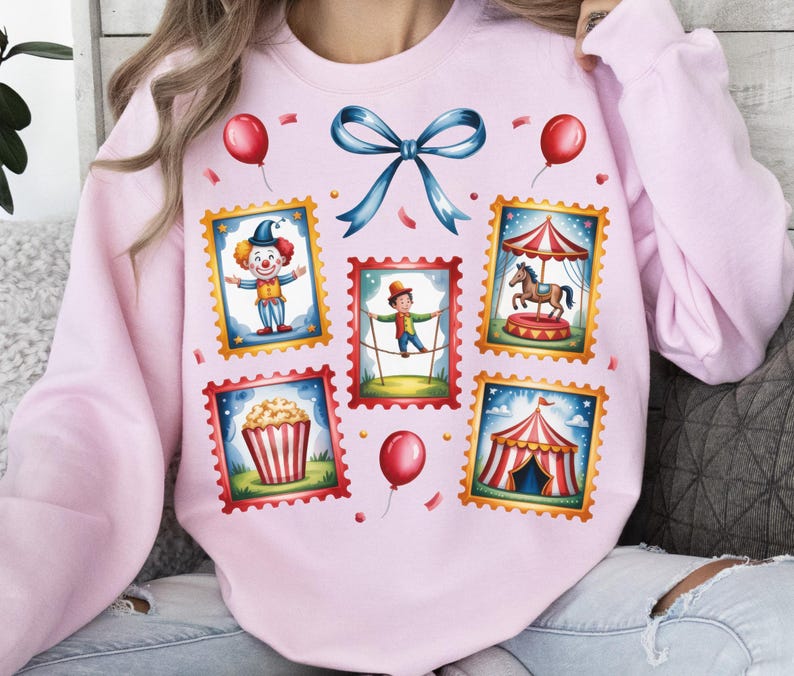 May include: A light pink sweatshirt featuring a circus-themed design. The design includes postage stamp-style illustrations of a clown, a tightrope walker, a carousel, popcorn, and a circus tent. Red balloons and a blue bow complete the design.