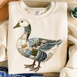 Può includere: Felpa color crema con un dettagliato design a patchwork di un'oca. L'oca ha una testa bianca, un becco verde e ali con toppe a motivi blu, verdi e marroni. La felpa ha un girocollo e maniche lunghe, perfetta per l'abbigliamento casual.