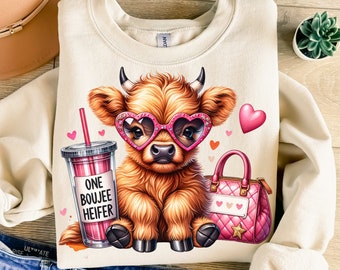 May include: A cream-colored sweatshirt with a cartoon illustration of a brown and white Highland cow wearing pink heart-shaped sunglasses. The cow is holding a pink purse and a pink and white cup with a straw that says "One Boujee Heifer".