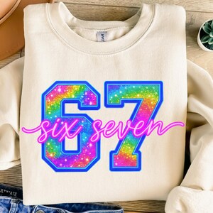 K&ouml;nnte beinhalten: Cremefarbenes Sweatshirt mit der Zahl "67" in einem Regenbogen-Glitzer-Design, umrandet in Blau. Die Worte "six seven" sind in rosa Schreibschrift geschrieben. Ein l&auml;ssiges, bequemes Oberteil.