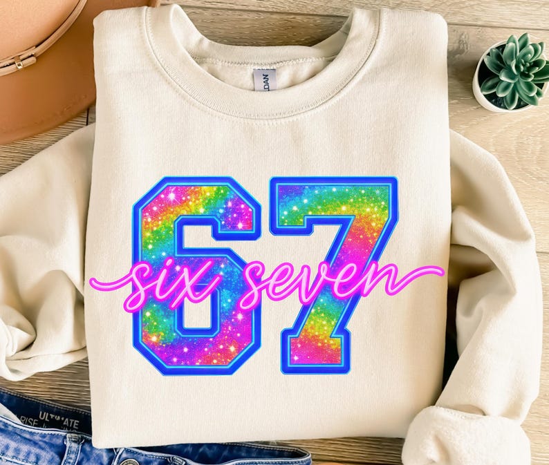 K&ouml;nnte beinhalten: Cremefarbenes Sweatshirt mit der Zahl "67" in einem Regenbogen-Glitzer-Design, umrandet in Blau. Die Worte "six seven" sind in rosa Schreibschrift geschrieben. Ein l&auml;ssiges, bequemes Oberteil.