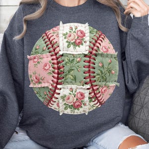 Może przedstawiać: Szara bluza z nadrukiem w postaci baseballu z kwiatowym wzorem. Baseball wykonany jest z różowych i zielonych kwiatowych łatek materiału, z czerwonymi szwami. Kwiatowy wzór zawiera różowe róże i zielone liście.