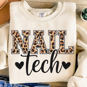 May include: Cream-colored sweatshirt with the words "NAIL tech" in black script and leopard print block letters. Two black hearts flank the word "tech". A small potted succulent is visible in the upper right corner.