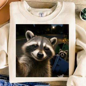 May include: Cream-colored sweatshirt featuring a raccoon selfie design. The image shows a raccoon taking a selfie with a trash can and another raccoon in the background. The design is printed on a soft, comfortable fabric.